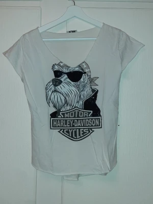 Vit t-shirt med tryck från LSK - Säljer en vit t-shirt från LSK "Motor Cycles Harley-Davidson" med ett stort tryck av en hund med solglasögon och bandana på bröstet. T-shirten har vid urringning och korta ärmar. Perfekt för dig som gillar coola motiv och streetwear-vibe. Storlek S/M.