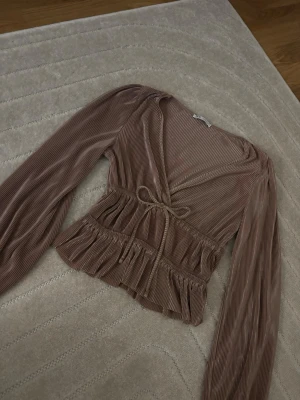 Champagne färgad plisserad blus från H&M - Snygg champagne färgad plisserad blus från H&M med långa ärmar och v-ringning. Blusen har ett knytband i midjan och volangdetaljer nedtill. Materialet är mjukt och glansigt, vilket ger en lyxig känsla. Perfekt för att skapa en trendig look.