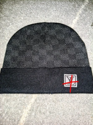 Mörkgrå mönstrad mössa med patch - Snygg mörkgrå mössa med diskret rutmönster och uppvikt kant. På framsidan sitter en fyrkantig patch med broderad detalj i svart, vitt och rött. Perfekt accessoar för kyliga dagar och passar till de flesta outfits.