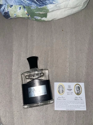 Creed Aventus Eau de Parfum 120ml - Creed Aventus är en lyxig parfym i en stilren, genomskinlig glasflaska med svart etikett och svart kork. Flaskan rymmer 120 ml och har märkets logga i silver. Aventus är känd för sin fräscha och fruktiga doftprofil och exklusiva känsla.
