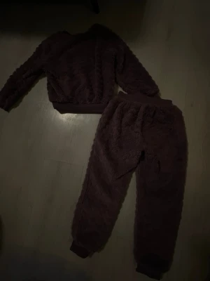 Mörklila  fluffigt loungewear-set - Mysigt tvådelat loungewear-set i mörklilla färg med fluffig textur. Setet består av en långärmad tröja och matchande byxor med resår i midjan och vid benslut. Perfekt för hemmamys och avslappnade dagar.