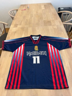 Maiden blå-röd t-shirt med nummer limited edition  - Snygg blå t-shirt från Maiden med röda och ljusblå ränder längs sidorna och ärmarna. Tryck med Eddie , Maiden-logga och siffran 11 både fram och bak. Klassisk sportig look med korta ärmar och v-ringad hals. Perfekt för dig som gillar unik streetwear.