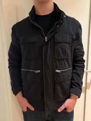 Svart vindjacka/fieldjacket från J.Lindeberg - Snygg svart vindjacka från J.Lindeberg med flera fickor framtill och dragkedjor. Jackan har hög krage, lång ärm och en liten logga på ena ärmen. Perfekt för dig som vill ha en stilren och funktionell jacka till vardags.