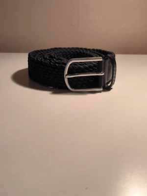 Svart flätat onewear bälte med silverfärgat spänne - Snyggt svart flätat onewear bälte med ett stilrent silverfärgat metallspänne. Bältet har en klassisk design och är gjort i ett syntetmaterial som ger en flexibel och bekväm passform. Perfekt för att lyfta vilken outfit som helst.