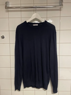 Mörkblå tröja från Cubus  - En stilren mörkblå tröja från Cubus med rund halsringning och långa ärmar. Tröjan har en enkel och klassisk design med ribbade muddar vid ärmslut och nederkant och är gjord i merinoull. Perfekt för en clean och avslappnad look.