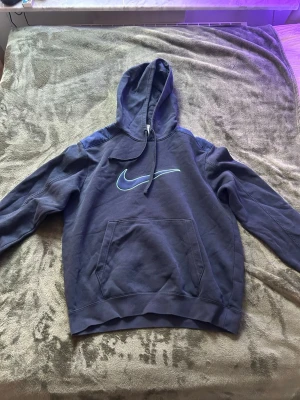 Mörkblå Nike hoodie med logga - Snygg mörkblå hoodie från Nike med stor ljusblå Swoosh-logga på bröstet. Klassisk känguruficka framtill och justerbar huva med dragsko. Tillverkad i mjukt bomullsmaterial som är perfekt för chill dagar eller streetwear-stil.