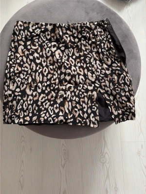 Leopardmönstrad kortkjol med slits - Säljer en snygg kortkjol med leopardmönster i svart, beige och bruna toner. Kjolen har en liten slits framtill som ger en extra detalj. Perfekt för dig som vill sticka ut med ett djurmönster i garderoben.