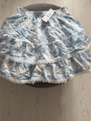 Blommig volangkjol från Gina Tricot - Supersöt kort kjol från Gina Tricot med blått blommönster på vit botten. Kjolen har volanger och spetsdetaljer längs kanterna samt elastisk midja för skön passform. Perfekt för en fräsch och somrig look!