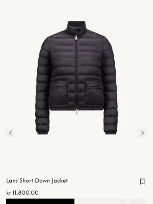 Svart dunjacka från Moncler - Snygg svart dunjacka från Moncler med kort, smal passform och hög krage. Jackan har två stora fickor med lock och knapp framtill, quiltad design och dragkedja. Perfekt för kalla vinterdagar och riktigt stilren look.