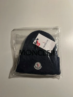 Mörkblå ribbstickad Moncler mössa - Snygg mörkblå ribbstickad mössa från Moncler med uppvikt kant och klassisk Moncler-logga framtill. Tillverkad i mjukt material som håller dig varm och ger en clean look. Perfekt accessoar för kyliga dagar. Kommer med en påse och prislapp. Prislappen har tyvärr åkit av. Inte använd en enda gång!😃