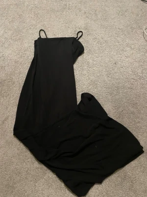 Svart långklänning med tunna band - En stilren svart långklänning med tunna axelband och rak passform. Klänningen har en öppning/slit nere vid ena benet. Banden är uppsydda på bilden då den har långa band men kommer såklart ta bort de innan den skickas.