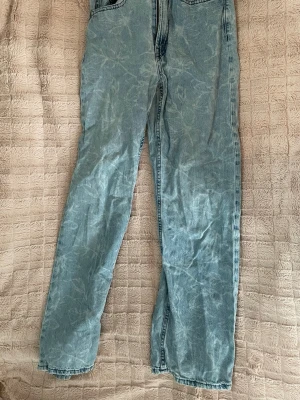 Ljusblå jeans med blomtryck  - Säljer ett par ljusblå jeans med acid wash-effekt och raka ben. Jeansen har klassisk femficksdesign och är tillverkade i denim. Perfekta för dig som gillar en avslappnad och trendig stil. Tyvärr finns ett litet hål baktill.
