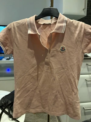 Beige pikétröja från Moncler - Säljer en beige pikétröja från Moncler med klassisk krage och knappar framtill. Tröjan har Moncler-logga på bröstet och vita detaljer på kragen. Perfekt för en clean och stilren look.