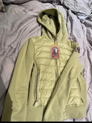  Parajumpers - Snygg ljusgrön hoodie/jacka från Parajumpers med quiltad front och mjuka ärmar. Dragkedja framtill, två fickor med dragkedja och en stor huva. Märkespatch på ärmen och ribbade muddar. Perfekt för dig som vill ha en sportig och trendig look. Storlek ys motsvarar xs. Lappar finns