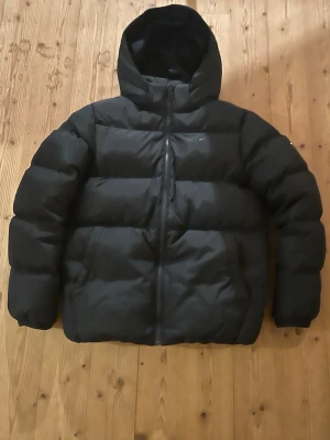 Svart pufferjacka Tommy Hilfiger - Snygg svart pufferjacka från Tommy Hilfiger med huva och dragkedja framtill. Jackan har en klassisk quiltad design och diskret logga på bröstet. Perfekt för kalla dagar och riktigt skön att bära. Modellen heter The New York Puffer.