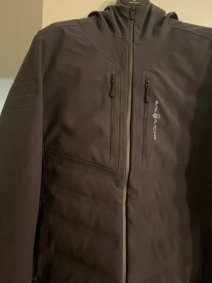 Svart vindjacka från Sail Racing - Snygg svart vindjacka från Sail Racing med två bröstfickor med dragkedja och logga på bröstet. Insidan har en cool gul vadderad detalj som sticker ut. Kvitto finns från sailracing butik vid NK innan dem gjorde om. Passar ej hänger bara i garderoben.