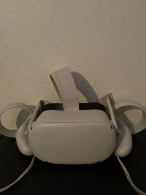 Oculus Quest 2 VR-headset med kontroller - Säljer ett Oculus Quest 2 VR-headset i vit plast med matchande justerbart huvudband och två handkontroller. Headsetet har en modern, rundad design och är trådlöst för maximal rörelsefrihet när du spelar VR-spel.