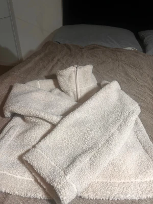 Vit teddyjacka med dragkedja - Mysig vit teddyjacka med hög krage och dragkedja framtill. Jackan har två sidofickor och är gjord i fluffigt, mjukt teddy-material som håller dig varm. Perfekt för kyliga dagar och ger en avslappnad, trendig look.