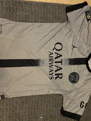 PSG Mbappé matchtröja grå - Snygg grå PSG matchtröja med svarta detaljer, Qatar Airways-tryck på framsidan och Mbappé 7 på ryggen. Tillverkad i lätt och ventilerande material, perfekt för fotboll eller träning. Ikoniskt PSG-märke på bröstet och Jordan-logga.