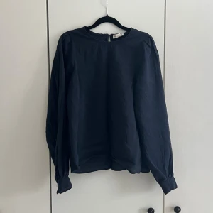 Mörkblå blus med rund hals - Stilren mörkblå blus från Lindex med rund halsringning och långa ärmar. Blusen har en rak passform och knäppning bak i nacken. Tillverkad i ett lätt, vävt material som ger ett snyggt fall. Perfekt att matcha med jeans eller kjol.