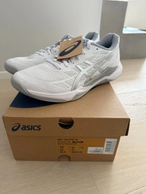  Asics GEL-Tactic 12 sneakers, oanvända  - Snygga vita Asics GEL-Tactic 12 sneakers med silverdetaljer och meshpaneler för bra ventilation. Storlek 42 (som 40 i vanliga skor). Oanvända och säljes pga ångrat köp. Passar för alla inomhussporter. Nypris ca 850 sek