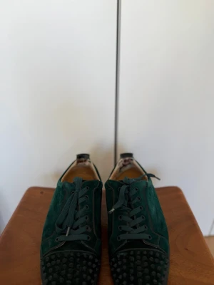 Christian Louboutin - Mörkgröna sneakers i mocka från Christian Louboutin med svarta nitar på tån och klassisk röd sula. Skorna har snörning och låg profil, vilket ger en edgy och unik look. Perfekta för dig som vill sticka ut med exklusiva detaljer och cool design.