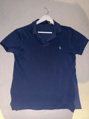 Oanvänd Marinblå pikétröja från Polo Ralph Lauren - Oanvänd Klassisk marinblå pikétröja från Polo Ralph Lauren med korta ärmar och den ikoniska broderade loggan på bröstet. Tröjan har krage och knappar vid halsen. Perfekt till jeans eller shorts för en clean och stilren look. Nypris är över 1500kr