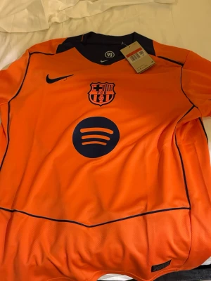 Orange FC Barcelona matchtröja Nike - Snygg orange FC Barcelona matchtröja från Nike med mörkblå detaljer. Tröjan har klubbmärke, Nike-logga och Spotify som sponsor framtill samt UNHCR-logga bak. Tillverkad i lätt och ventilerande material, perfekt för fotboll eller träning.