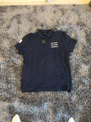 Mörkblå Peak Performance piké - Snygg mörkblå pikétröja från Peak Performance med vit logotyp på bröstet. Klassisk krage och knappar framtill. Tillverkad i mjuk bomull som känns skön mot huden. Perfekt för en clean och sportig stil.ordi pris 499kr