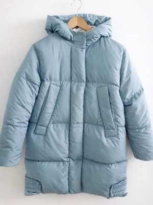 Ljusblå lång dunjacka med huva - Snygg ljusblå dunjacka med huva och långa ärmar. Jackan har två snedställda fickor framtill och quiltad design som ger en puffig look. Perfekt för kalla dagar och har en stilren, minimalistisk känsla.