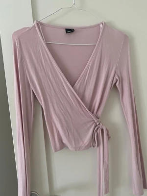 Ljusrosa omlottopp från Gina Tricot - Supersöt ljusrosa omlottopp från Gina Tricot med långa ärmar och knyt i sidan. Tunn och mjuk, perfekt till jeans eller kjol. Snygg v-ringning och smickrande passform.