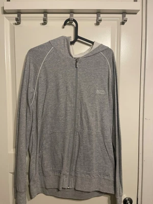 Grå zip-hoodie från BOSS - Snygg grå hoodie med dragkedja från BOSS. Den har huva, vita detaljer längs ärmarna och två fickor framtill. Perfekt för en avslappnad stil och enkel att matcha med andra plagg. Materialet känns mjukt och bekvämt.
