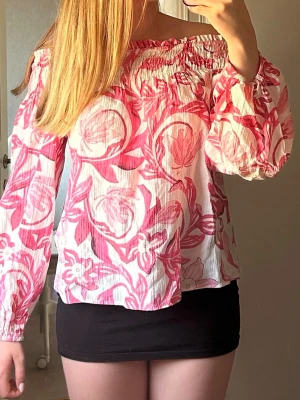 Offshoulder blommig sommrig blus  - Säljer en söt offshoulder blus med rosa och vita blommönster. Blusen har långa ärmar med resår vid axlar och ärmslut. Materialet är lätt och luftigt, perfekt för sommaren.
