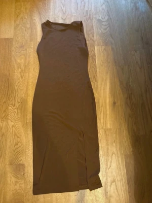 Brun långklänning med slits - En stilren brun långklänning i figurnära modell med ärmlös design och rund halsringning. Klänningen har en diskret slits framtill som ger en snygg detalj. Perfekt för dig som gillar minimalistisk stil.