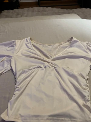 Vit omlottopp med v-ringning - Snygg vit topp med omlottdesign och v-ringning. Långa ärmar och lätt rynkade sidor ger en modern och fräsch look. Perfekt att styla med jeans eller kjol för en trendig outfit.