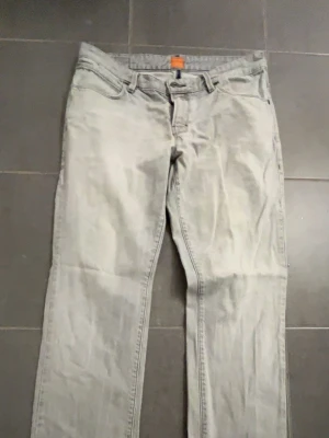 Ljusgrå Hugo boss jeans - Säljer ett par ljusgrå Hugo boss jeans med klassisk femficksdesign och raka ben. Jeansen har en orange detalj på linningen och är tillverkade i denim. Perfekt för dig som gillar en enkel och stilren look. Storlek 33:32, pris kan diskuteras vid snabb affär!