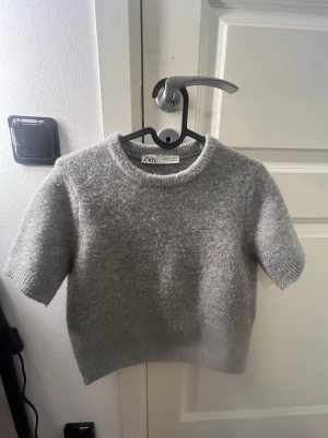 Grå stickad t-shirt från Zara - En grå stickad t-shirt från Zara med korta ärmar och rund halsringning. Tröjan har en mjuk och fluffig känsla, ribbstickade detaljer vid hals, ärmslut och nederkant. Perfekt för lager-på-lager och en chill look.