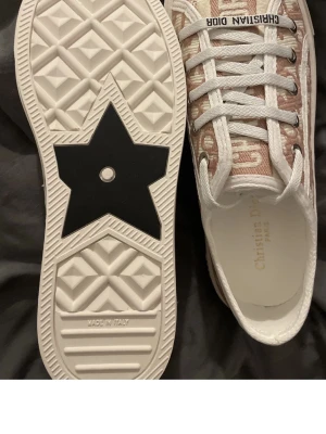 Christian Dior sneakers med monogram - Snygga låga sneakers från Christian Dior med beige och vitt monogrammönster, vita snören och tjock vit sula med svart stjärndetalj undertill. Ovandelen är i textil och sulan i gummi. Märkeslogga på plösen och sidan.