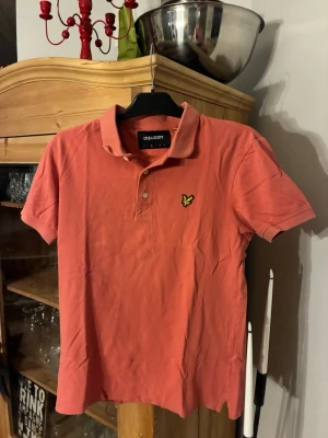 Korallröd pikétröja från Lyle & Scott - Snygg korallröd pikétröja från Lyle & Scott med klassisk krage och korta ärmar. Tröjan har två knappar framtill och den ikoniska gula loggan broderad på bröstet. Tillverkad i mjuk bomull för en bekväm känsla. Använd ett fåtal gånger och har storleken M