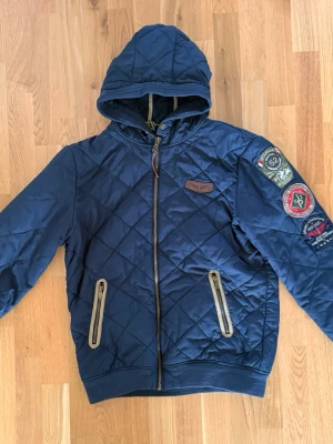 Marinblå vindjacka från Jean Paul - Snygg marinblå vindjacka från Jean Paul med quiltat mönster, huva och dragkedja framtill. Jackan har två fickor med dragkedja och coola patchar på ena ärmen. Perfekt för blåsiga dagar och streetstyle.