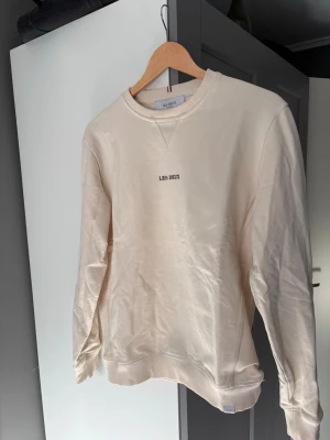 Beige sweatshirt från Les Deux - Säljer en stilren beige sweatshirt från Les Deux med rund halsringning och diskret logga broderad på bröstet. Tröjan har ribbade muddar vid ärmslut och nederkant samt en klassisk relaxed fit. Perfekt för dig som gillar minimalistisk och clean stil.