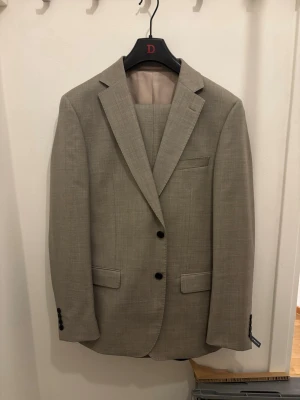Beige kostym  - Snygg beige kostym med klassisk kavaj och matchande byxor. Kavajen har två knappar framtill, smala slag och fickor med lock. Perfekt för dig som vill ha en stilren och tidlös look. Använd 1 gång, har nån liten fläck som går att tvätta bort. Köp för 2999kr