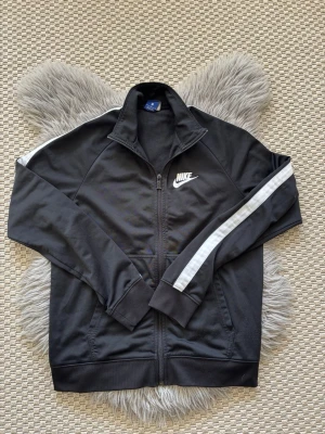 Svart Nike träningsjacka med vit rand - Snygg svart träningsjacka från Nike med vit logga på bröstet och vita ränder längs ärmarna. Jackan har dragkedja framtill, hög krage och muddar vid ärmslut och nederkant. Perfekt för träning eller chill.