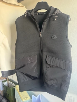 Svart Moncler väst med huva - Snygg svart väst från Moncler med huva och dragkedja framtill. Västen har stora fickor med lock i blankt material och Moncler-logga på bröstet. Perfekt för lager på lager och streetwear-stil. Materialmix av textil och nylon på fickorna.