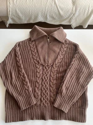 Brun kabelstickad Gant tröja - Säljer en brun kabelstickad tröja med hög krage och halvzip med ringdetalj. Tröjan har breda ribbade muddar och ett snyggt flätmönster framtill och på ärmarna. Perfekt för dig som gillar en avslappnad men trendig look.