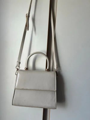 Vit fyrkantig handväska med axelrem - Stilren vit handväska i fyrkantig modell med justerbar axelrem och ett litet handtag upptill. Väskan har guldfärgade detaljer och är tillverkad i glansigt konstläder. Perfekt för dig som gillar minimalistisk och trendig design.kan användas utan axelband för en finare och mer elegant tillfälle.