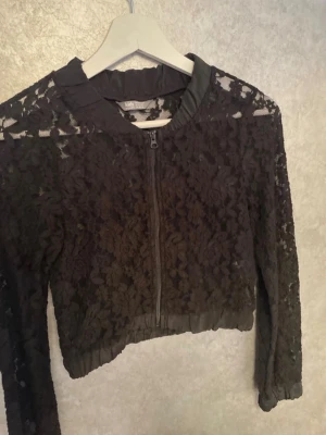 Svart spetsjacka med dragkedja - Snygg svart spetsjacka från Kids med genombrutet blommönster och dragkedja framtill. Kortare modell med långa ärmar och ribbad kant nedtill. Perfekt för att lyfta en outfit med en trendig touch.