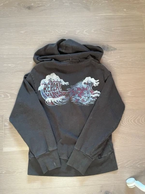 Svart hoodie från Replay med tryck - Säljer en svart hoodie från Replay med ett stort våg- och drakmotiv i vitt, rött och blått på ryggen. Hoodien har dragkedja framtill, huva och breda muddar vid ärmar och nederkant. Perfekt för dig som gillar streetwear och coola detaljer.