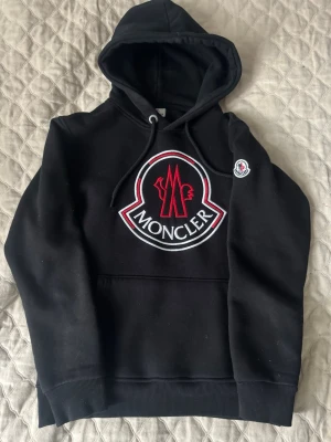 Svart Moncler hoodie  - Svart hoodie från Moncler med stor broderad logga i rött och vitt på bröstet samt en mindre logotyp på ärmen. Klassisk huva med dragsko och magficka. Mjuk bomullsblandning och ribbade muddar vid ärmslut och nederkant. 