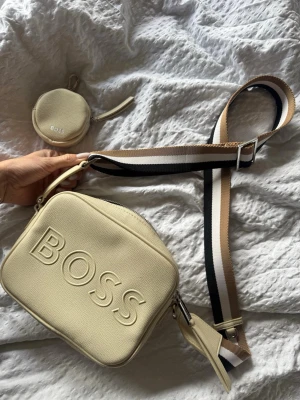 Beige handväska från BOSS - Stilren beige handväska från BOSS i strukturerat konstläder med matchande rund miniväska. Väskan har bred, justerbar axelrem i beige, vitt och svart. Stor BOSS-logga framtill och dragkedja upptill. Perfekt accessoar för en trendig look. Säljs för 1600-2000kr helt nya!